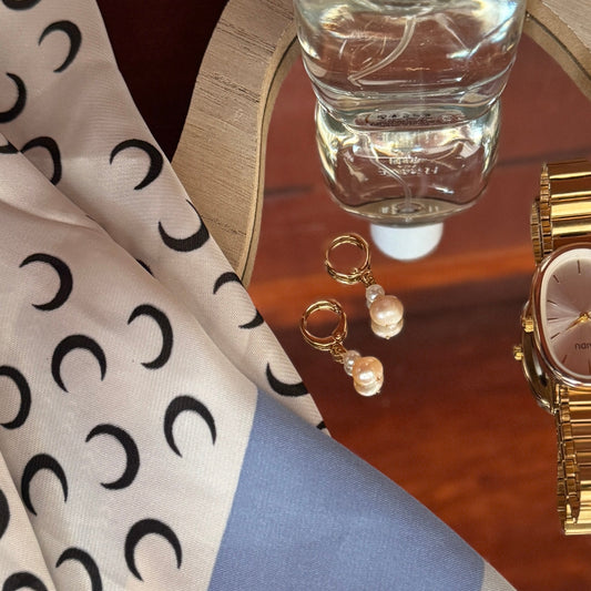 Satin scarf with moon pattern, bottle of hand sanitizer, watch, and earrings on a wooden surface.