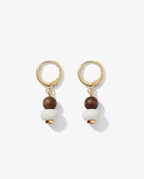 A pair of gold hoop drop earrings featuring a small dark brown wooden bead, a larger white spherical bead, and a tiny red accent bead at the bottom, displayed on a clean white background.